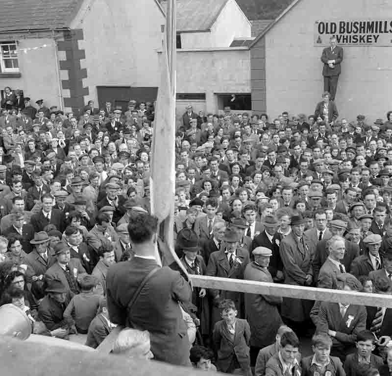 Eine Schwarz-Weiß-Fotografie aus einer erhöhten Perspektive zeigt eine große Menschenmenge in Irland, die sich im Freien zu einer Kundgebung zusammengefunden hat. Im Hintergrund sind einfache, helle Gebäude zu sehen. An der Wand eines Gebäudes auf der rechten Seite ist ein Schild mit der Aufschrift 'OLD BUSHMILLS WHISKEY' angebracht.