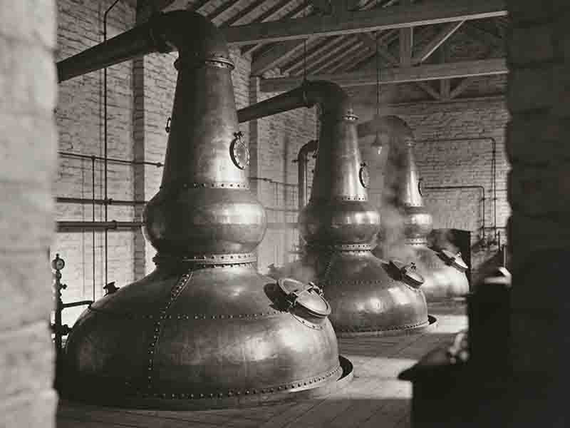 Schwarz-Weiß-Aufnahme von drei traditionellen kupfernen Pot Stills in einer historischen schottischen Destillerie mit Backsteinwänden und Holzbalkendecke.